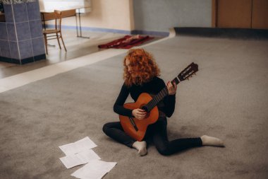 Kızıl saçlı bir kadın halı döşemelere oturmuş, elinde bir gitar tutuyor ve müzik çalışıyor. Gevşek notalar etrafını sarmış, dingin ve odaklanmış bir atmosfer yaratmış..