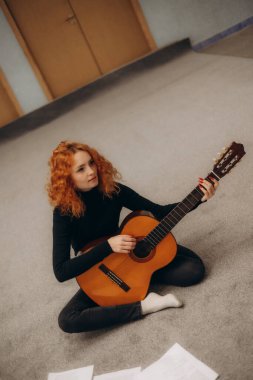 Kızıl saçlı bir kadın iç mekanda akustik gitar çalarak bacak bacak üstüne atmış oturuyor. Huzurlu ifadesi ve müzik etkinliği, rahat ve rahat bir ortamda yaratıcılık, odaklanma ve zevk göstergesi..