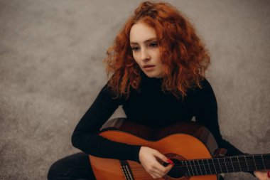 Canlı kızıl saçlı genç bir kadın evde otururken akustik gitar çalıyor, yaratıcılık ve içgözlem hissi yayıyor..