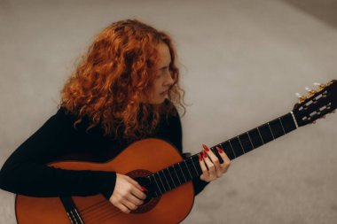 Kıvırcık kızıl saçlı ve tırnaklarını boyayan genç bir kadın akustik gitar çalıyor, sakin ve minimalist bir ortamda müziğine odaklanıyor..
