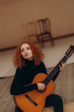Siyah kıyafetli kızıl saçlı bir kadın evde akustik gitar çalıyor, sıcak ve sakin bir atmosferi var. Kompozisyon, basit, sade bir iç mekanda sanatçılık ve tefekküre vurgu yapıyor..