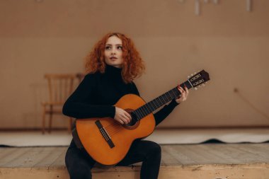 Kızıl saçlı bir kadın klasik akustik gitar çalar, siyah bir kıyafet giyer, müziğe olan tutkusunu ifade eder. Dekorun minimalist ve sanatsal bir atmosferi var..