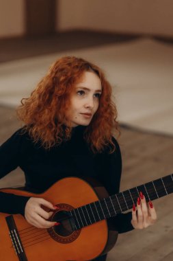 Kızıl saçlı bir kadın evde akustik bir gitarla pratik yapıyor. Tutku ve sükunet taşıyor..