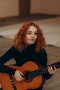 Koyu renk elbiseli sakin kızıl saçlı bir kadın içeride akustik gitar çalıyor. Ahşap döşeme ve yumuşak tonlar barışçıl ve uyumlu bir atmosfer yaratır..