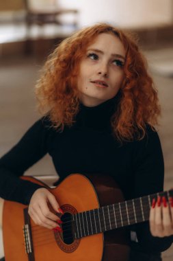 Kapalı mekanda oturan kızıl saçlı bir kadın, düşünceli bir şekilde akustik gitar çalıyor, sanatsal ve huzurlu bir atmosfer yaratıyor..