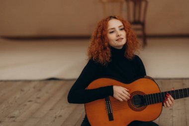 Kızıl kıvırcık saçlı, akustik gitar çalan genç bir kadın minimalist kapalı bir ortamda otururken sıcak ve sanatsal bir hava veriyor..