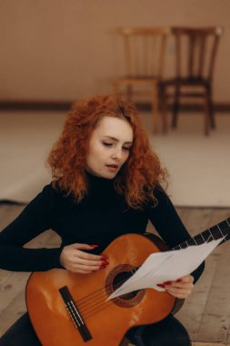 Akustik gitar çalan ve sıcak, minimalist bir ortamda nota okuyan kızıl saçlı genç bir kadın. Yaratıcılık, müzik tutkusu ve sanatsal ifade anı yakalamak, öğrenme ve kararlılığı yansıtır..