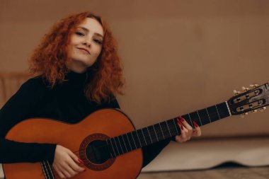 Canlı kızıl saçlı bir kadın, siyah bir bluz giyerken akustik gitar çalar, sakin ve sanatsal bir atmosfer yaratır..