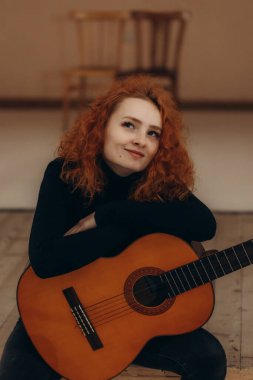 Evde oturan akustik gitarlı kızıl saçlı kadın neşeli ve yaratıcı bir ortam sergiliyor. Sıcak ortam, sessiz anın tadını çıkarırken kişinin sanatsal ve dışavurumsal doğasını vurguluyor..