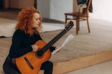 Evde nota okurken, elinde gitar tutan kızıl saçlı genç bir kadın yaratıcılık, odak ve sanatçılık aktarıyor. Dekor, müzikal ifade ve pratik için mükemmel sıcak ve ilham verici bir ortam sergiliyor..
