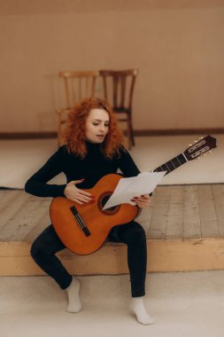 Kızıl saçlı genç bir kadın, müzik kağıtlarını tutarken akustik gitar çalıyor, kapalı bir mekanda oturuyor. Sahne yaratıcılığı, odağı ve müzikal ilhamı çağrıştırıyor.