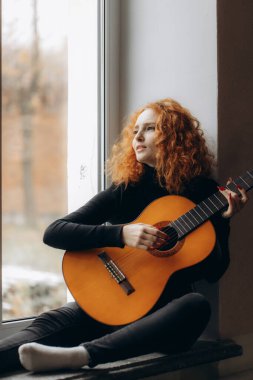 Pencere kenarında oturan kızıl saçlı bir kadın akustik gitarını çalar, sakin ve rahat bir ortamda düşünceli ve yaratıcı bir enerji yayar..