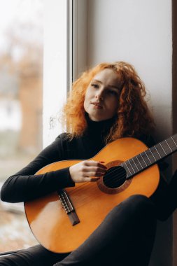 Kıvırcık saçlı genç bir kadın pencerenin kenarında oturur. Elinde sakin ve düşünceli bir gitar tutar. Sıcak ve içe dönük bir atmosfer yaratır..