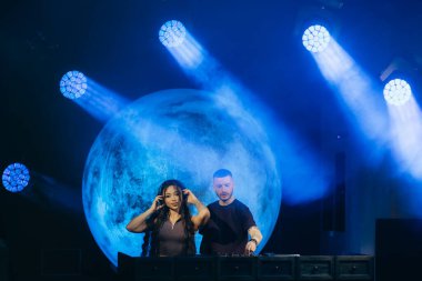 DJ 'ler bir müzik etkinliğinde canlı performans sergiliyorlar, büyük dairesel dekoratif arka planla mavi tiyatro ışıklandırmasıyla aydınlatılıyorlar..
