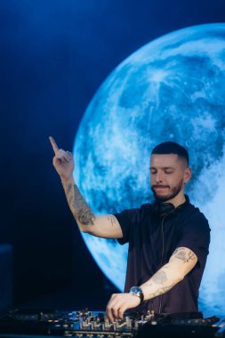 Bir DJ, bir güvertede müziği karıştırırken yoğun bir şekilde odaklanır, büyük mavi bir arka planla, kulübün atmosferini yakalar ve bir müzik performansının sanatsal havasını yakalar..