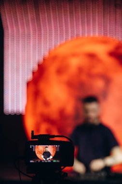 Canlı yayında bir DJ 'i kaydeden bir kameranın canlı görüntüsü. Parlak kırmızı dairesel arkaplan ve bulanık odak sanatsal ve enerjik ortamı vurgular. Etkinlik prodüksiyonu ya da eğlence temaları için mükemmel.