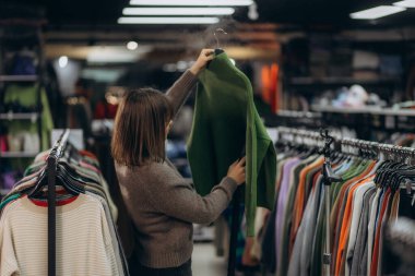 Bir kadın, renkli giysi ve askılarla çevrili iyi ışıklandırılmış bir giyim mağazasında, modern bir perakende mağazasında moda alışveriş deneyimini vurgulayan yeşil bir kazak seçiyor..