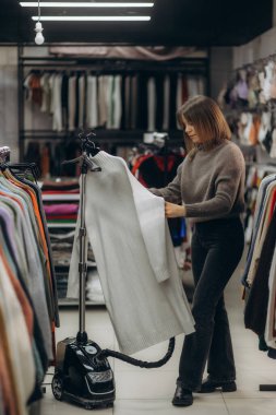 Bir kadın giysi buharlı tenceresini bir giyim mağazasında, giysi mağazasında takılıyor, giysi ve satış için giysilerin korunması ve hazırlanması sürecini gösteriyor..