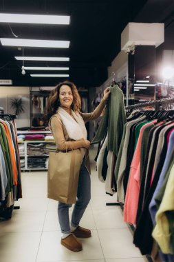 Neşeli bir kadın kıyafet alışverişi yapıyor, alışveriş çantası taşıyor ve renkli moda eşyalarıyla çevrili modern bir perakende mağazasında eğleniyor..
