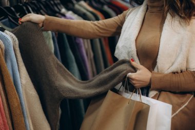 Şık bir kadın, bir giyim mağazasında elinde alışveriş torbalarıyla örgü kıyafetlerini inceliyor, sonbahar moda seçimlerini vurguluyor..