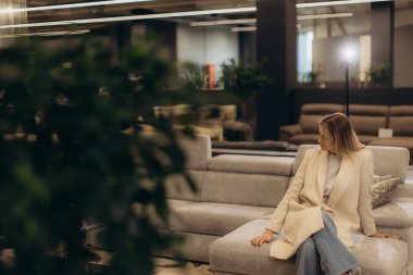 Sofistike bir ceket giyen bir kadın, şık dekore edilmiş bir mobilya galerisinde gri bir kanepeye rahatça oturur ve rahatlama ve lüks bir hayat sunar..