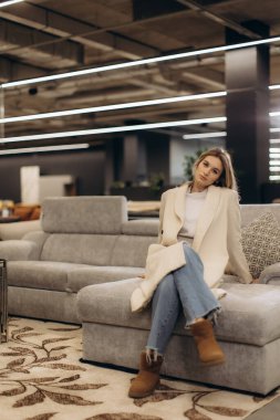 Beyaz önlük ve kot pantolon giyen genç bir kadın şık bir şekilde tasarlanmış, rahat dekorlu ve hafif ışıklandırmalı modern bir koltukta oturuyor..