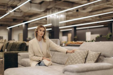 Zarif giyimli bir kadın modern tasarımlar ve nötr tonlarla çevrili çağdaş bir mobilya galerisinde gri bir koltukta oturur..