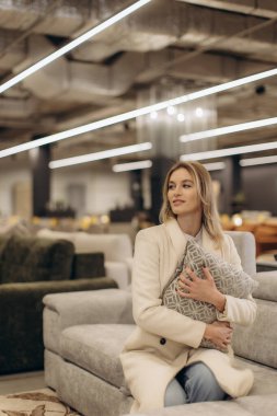 Modern bir mobilya galerisinde dekoratif bir yastık tutarken şık giyimli bir kadın koltukta dinleniyor. Sahne, perakende ortamında konfor ve zarif iç tasarım sergiliyor..