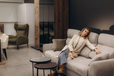 Modern bir koltukta oturan bir kadın güzel tasarlanmış bir yaşam alanında koltuklar ve masalar dahil çağdaş mobilyalarla ev dekorasyonunda rahatlık ve zarafet hissi uyandırıyor..