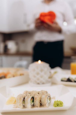 Mum ışığında romantik bir atmosferi tasvir eden bir suşi tabağının yakın plan görüntüsü. Bulanık bir arka plan, birinin kırmızı bir hediye tuttuğunu gösteriyor, samimi havayı güçlendiriyor..