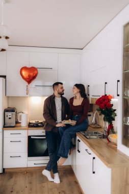 Bir çift mutfakta samimi bir anı paylaşıyor, kalp şeklinde bir balon ve gül buketi, aşkı ve kutlamaları simgeliyor..