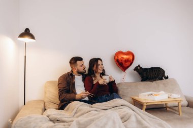 Rahat bir çift rahat bir kanepede oturuyor, yanında bir kedi ve kalp şeklinde bir balon var, sıcak ve davetkar bir evde kahve ve atıştırmalıklarla vakit geçiriyorlar..