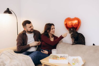 Mutlu bir çift kanepede oturup suşinin tadını çıkarıyor, yanında bir balon ve kedileri var. Sahne, sevgi, birliktelik ve sıcaklığı, arkadaşlığı lezzetli yemekler ve sıcak bir atmosferle kutluyor..