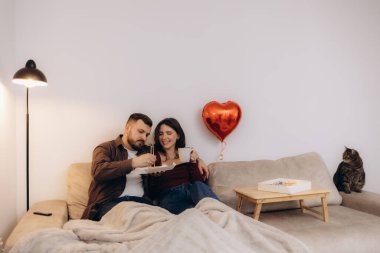 Kedi ve kalp şeklinde bir balon eşliğinde rahat bir kanepede dinlenen bir çift, sevgi ve rahatlık ortamı yaratıyor..