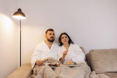 Kanepede rahat rahat oturan, bornoz giyen ve birlikte atıştırmanın tadını çıkaran bir çift. Ayarlar rahatlama, bağ kurma ve rahat bir ev ortamında huzurlu bir an olduğunu gösteriyor..