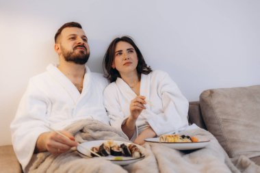 Beyaz bornozlu büyüleyici bir çift rahat bir anı paylaşıyor rahat bir kanepede suşi yiyorlar. Rahatlığın, romantizmin ve konforlu bir yemeğin mükemmel tasviri..