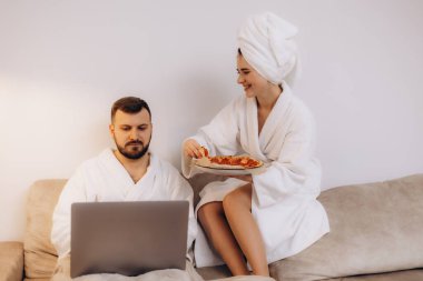 Bornozlu bir çift evde rahat bir anın tadını çıkarıyor. Kadın ona pizza ikram ederken, adam dizüstü bilgisayarda çalışıyor. İçerde rahatlık ve beraberlik sahnesi sergiliyor..