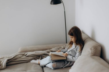 Genç bir kız kanepede rahat bir şekilde oturur, bir kitap okumaya dalmış, basit ve rahat bir oturma odası ve hafif ışıklandırmayla çevrili..
