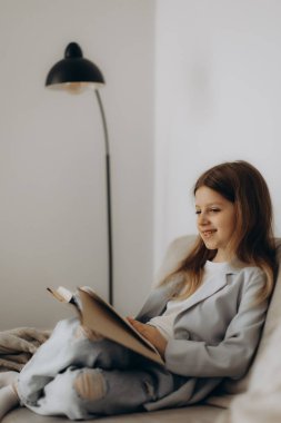 Genç bir kız kanepede oturur, rahat bir ortamda kitabının tadını çıkarırken gülümser. Huzurlu atmosfer ve doğal ışıklandırma bir dinlenme ve rahatlama anına katkıda bulunur..