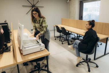 Modern ofis ortamında iki genç kadın, biri yazıcı kullanıyor ve biri de yakınlarda oturuyor..