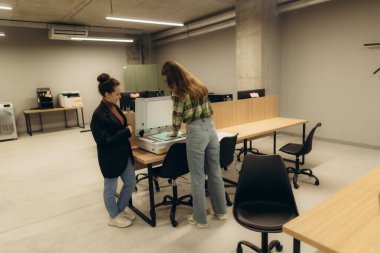 Çağdaş ofis ortamında tarayıcının yanında çalışan iki kadın profesyonel işbirliği sergiliyor. Sahnede takım çalışması, modern ofis ekipmanları ve iyi tasarlanmış bir çalışma alanı yer alıyor..