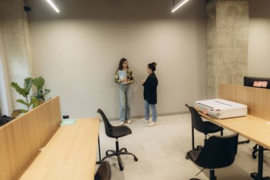 Masalar ve yeşilliklerle sohbet eden iki kadının bulunduğu sıcak ışıklandırılmış çağdaş bir ofis, etkileşim ve işbirliğini teşvik eden profesyonel ama davetkar bir alan yaratıyor..