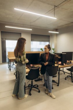 Modern bir ofiste bilgisayar masalarının yanında dururken sohbet eden iki meslektaş. Bu iş yeri senaryosunda şık iç tasarım ve profesyonel işbirliği vurgulanmış olup, takım çalışması kavramlarını göstermektedir.