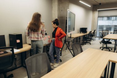 Modern bir ofis ortamında birlikte çalışan kadınlar ekipmanın yakınındaki görevleri tartışıyorlar. Çalışma alanı, temiz çizgiler, organize masalar ve yaratıcılık ve takım çalışmasının göstergesi olan profesyonel bir çevreye sahiptir..