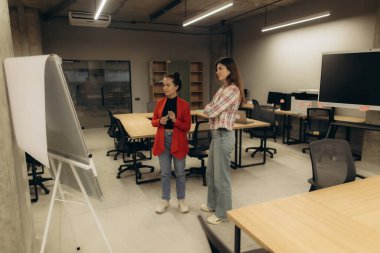 İki profesyonel kadın modern bir ofiste, beyaz tahtada fikirleri tartışıyor. Çalışma alanı çağdaş, masalar, etkileşimli bir ekran ve açık, üretken bir çevredir. İşbirliği ve yaratıcılık vurgulandı.