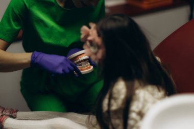 Yeşil üniformalı bir diş hekimi klinik ortamında diş modelini kullanan genç bir kıza uygun ağız temizliği uygulamasını gösterir..