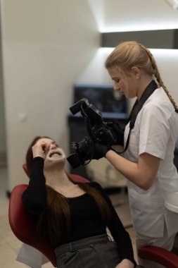 Profesyonel diş sağlığı kliniği sahnesi, bir dişçinin tıbbi belgeler ve tedavi süreci değerlendirmesi için dişlerinin fotoğrafını çekmesini, diş bakımı ve muayene teknolojilerini vurgulamasını göstermektedir..