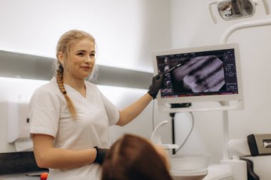 Beyaz üniformalı kadın dişçi monitörde diş röntgenlerini hastaya gösteriyor. Modern diş sağlığı teknolojisi ve hasta etkileşimini temiz ve profesyonel bir tıbbi ortamda gösteriyor..