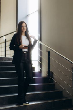 Kendine güvenen genç bir kadın modern bir iş ortamında resmi olarak ofis merdivenlerinden elinde akıllı bir telefonla iniyor. Sahne profesyonellik, odak ve çağdaş çalışma kültürünü aktarıyor.