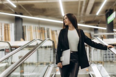 İş kıyafeti giymiş genç profesyonel bir kadın modern bir alışveriş merkezinin içinde yürüyen merdiven kullanıyor. Kendine güveni ve dikkati tam..